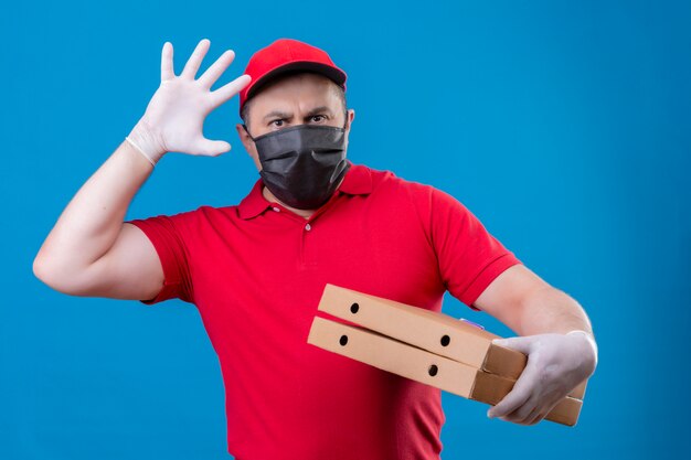 Fattorino che indossa l'uniforme rossa e cappuccio in scatole protettive facciali che tengono le scatole di pizza con la mano e la palma sollevate con l'espressione arrabbiata che controlla isolante