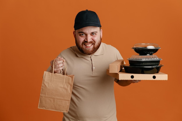 Fattorino dipendente in berretto nero e maglietta vuota uniforme che tiene contenitori per alimenti con sacchetto di carta guardando la fotocamera felice e contento sorridente allegramente in piedi su sfondo arancione