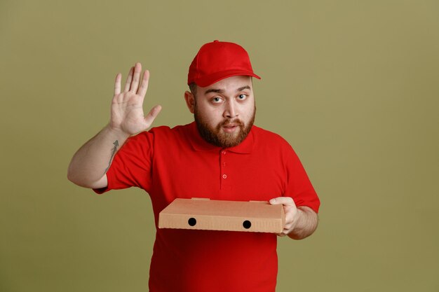 Fattorino dipendente in berretto rosso maglietta vuota uniforme che tiene scatola per pizza guardando la fotocamera felice e positivo agitando con la mano gesto di saluto in piedi su sfondo verde
