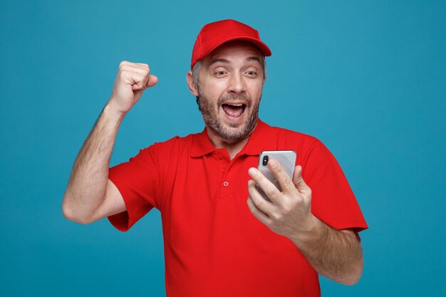 Fattorino dipendente in berretto rosso maglietta vuota uniforme che tiene smartphone guardando lo schermo pazzo felice ed eccitato pugno di serraggio gioendo del suo successo in piedi su sfondo blu