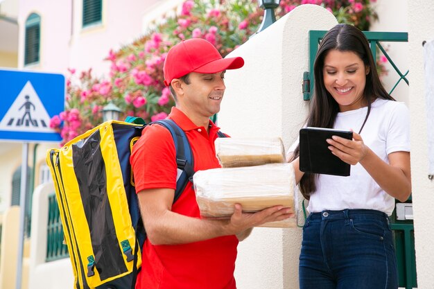 Fattorino felice in piedi vicino al cliente con tablet. Postino professionista in scatole di contenimento uniformi rosse e consegna dell'ordine. Cliente femminile grazioso che ottiene i pacchi. Servizio di consegna e concetto di posta