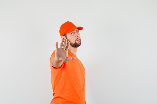 Fattorino in maglietta arancione, berretto che mostra il gesto di arresto e sembra esausto, vista frontale.