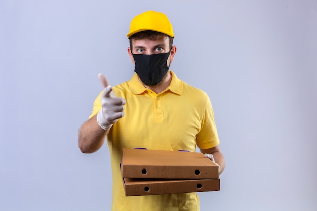 fattorino in maglietta polo gialla e berretto che indossa la maschera protettiva nera in possesso di scatole per pizza che punta il dito indice verso l'espressione della fotocamera come fare domanda su bianco isolato