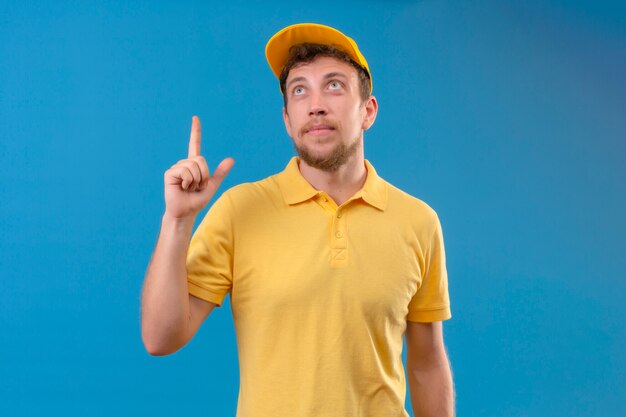 fattorino in maglietta polo gialla e berretto guardando fiducioso puntando con il dito in alto e alzando lo sguardo in piedi sul blu isolato