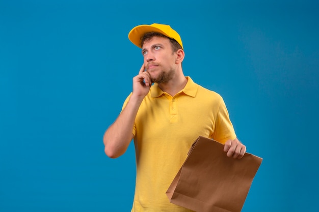 fattorino in maglietta polo gialla e berretto tenendo il pacchetto di carta in piedi con la mano sul mento alzando lo sguardo pensando pensieroso guardare sul blu isolato