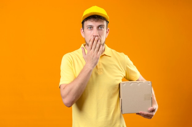 fattorino in maglietta polo gialla e berretto tenendo il pacchetto scatola scioccato coning bocca con la mano in piedi sul arancione isolato