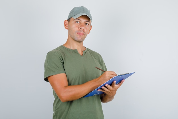 Fattorino prendendo nota negli appunti in t-shirt verde militare, berretto, vista frontale.