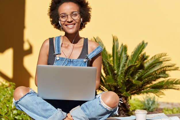 Felice adolescente afroamericano attraente con espressione positiva, posa nella posa del loto, utilizza il computer portatile per ascoltare musica e lavorare come freelance