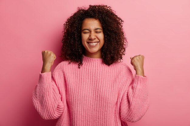 Felice adolescente dalla pelle scura solleva i pugni chiusi, celebra la vittoria e il successo, ha ottenuto il desiderio desiderabile, sorride ampiamente, vestito con un maglione lavorato a maglia, isolato sul muro rosa.