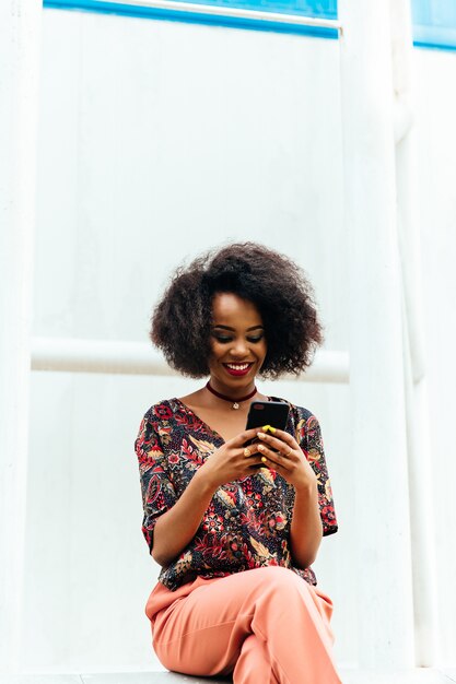 Felice affascinante donna afro-americana, guardando lo schermo smartphone, sorridendo allegramente