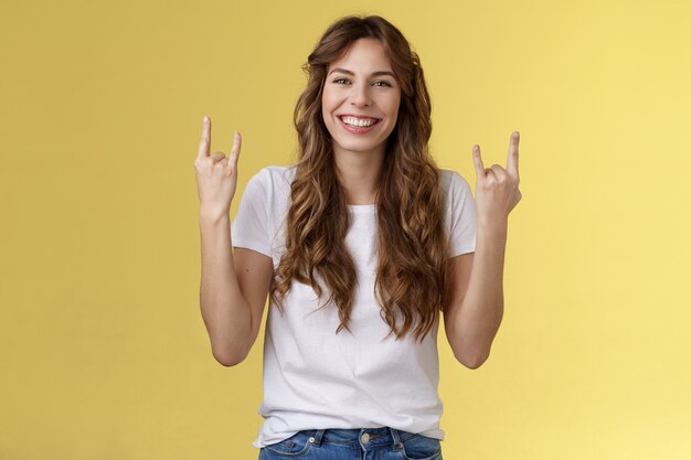 Felice allegro femminile caucasica ragazza riccia acconciatura spettacolo rock-n-roll heavy metal gesto sorridente ampiamente goditi l'incredibile atmosfera da concerto stare in piedi sfondo giallo con gioia tifo soddisfatto