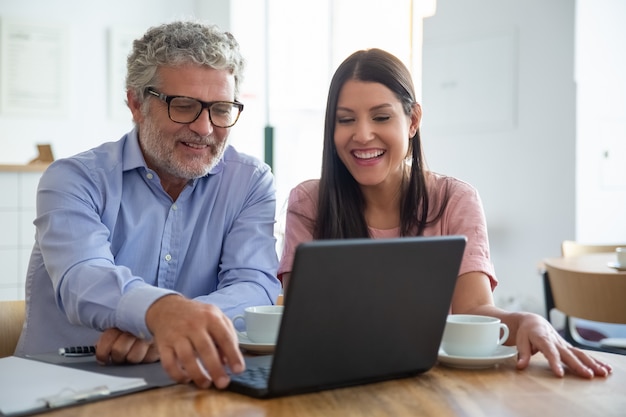 Felice allegro uomo maturo e giovane donna seduta al computer portatile aperto, guardando il display, guardando il contenuto davanti a una tazza di caffè e ridendo