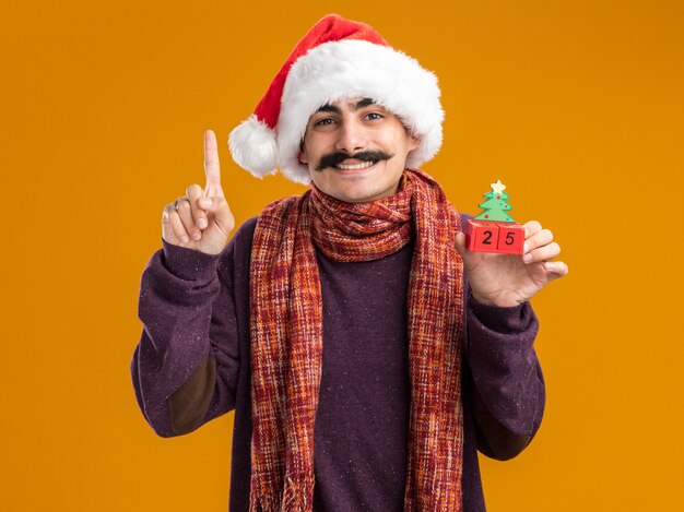 Felice baffuto uomo che indossa il natale santa hat con calda sciarpa intorno al collo tenendo i cubi giocattolo con data venticinque sorridendo allegramente mostrando il dito indice in piedi su sfondo arancione