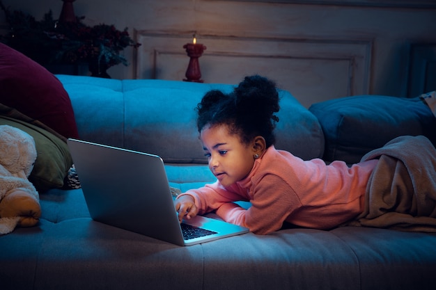 Felice bambina afro-americana durante la videochiamata con laptop e dispositivi domestici, sembra felice