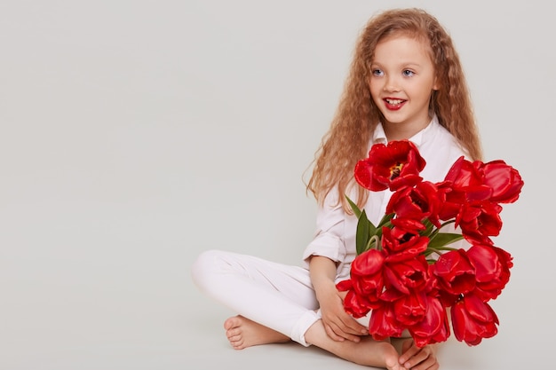 Felice bambina bionda che guarda lontano con un sorriso a trentadue denti e positiva espressione facciale, seduto sul pavimento, tenendo il mazzo di tulipani rossi