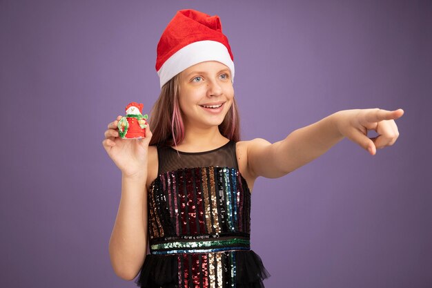 Felice bambina in abito da festa glitter e cappello da Babbo Natale che mostra il giocattolo di Natale che guarda da parte puntando con il dito indice a qualcosa sul lato in piedi su sfondo viola