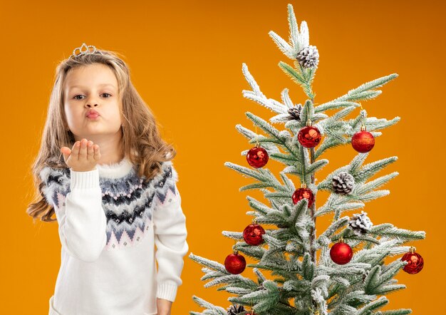 Felice bambina in piedi vicino albero di Natale che indossa tiara con ghirlanda sul collo che mostra gesto di bacio isolato su sfondo arancione