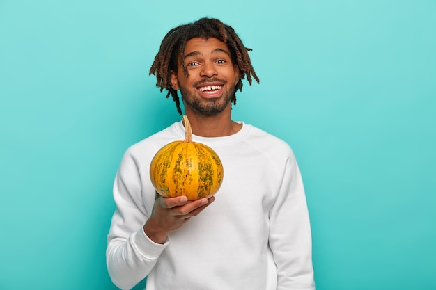 Felice bel giovane ragazzo di razza mista con dreadlocks, tiene poca zucca gialla matura, andando a fare insalata di dieta, vestito in abbigliamento casual