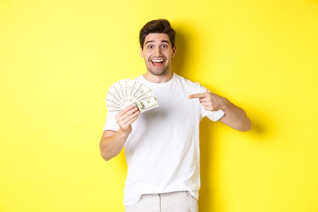 Felice bel ragazzo puntare il dito contro il denaro, il concetto di credito e prestito, in piedi su sfondo giallo.