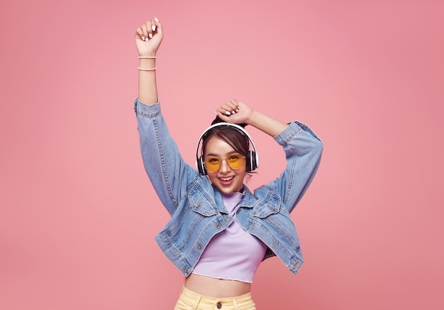 Felice bella donna asiatica adolescente in occhiali gialli che ascolta musica in cuffia e balla sulla parete rosa.