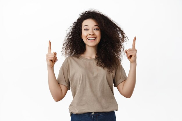 Felice bella donna che punta il dito verso l'alto, sorridente denti bianchi e mostrando pubblicità in cima, dimostrando lo spazio vuoto per banner, in piedi sullo sfondo dello studio