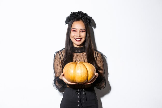 Felice bella donna in abito di pizzo nero godendo le vacanze di halloween, sorridendo alla telecamera e tenendo la zucca, in piedi su sfondo bianco.