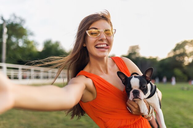 Felice bella donna parco facendo selfie foto, tenendo in mano il cane boston terrier, sorridente stato d'animo positivo, stile estivo alla moda, indossando abito arancione, occhiali da sole, giocando con animali, divertendosi