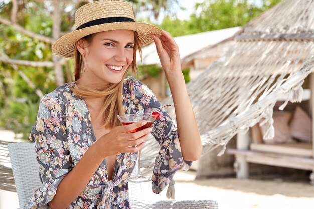 Felice bella donna rilassata si diverte all'aperto, indossa cappello e camicia di paglia, beve cocktail freschi, si siede su una sedia vicino all'amaca, ricrea in un paese tropicale. La donna allegra ha festa in spiaggia