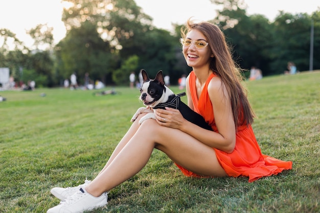 Felice bella donna seduta sull'erba nel parco estivo, tenendo in mano il cane boston terrier, sorridente stato d'animo positivo, indossa un abito arancione, stile alla moda, gambe sottili, scarpe da ginnastica, giocando con l'animale domestico