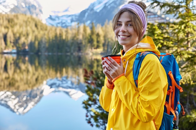 Felice bella donna viaggiatrice trascorre il tempo libero in località di montagna, beve caffè dalla tazza usa e getta