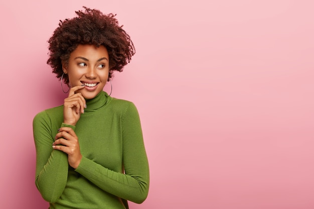 Felice bella femmina con i capelli ricci luminosi, sorride ampiamente, mostra i denti bianchi, tiene le mani vicino alla bocca, indossa dolcevita verde casual, guarda da parte, isolato sul muro rosa, spazio vuoto