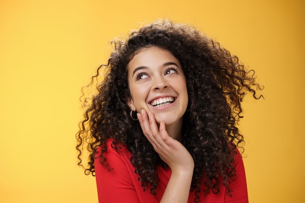 Felice bella giovane donna positiva con i capelli ricci in camicetta rossa che ride sciocco e spensierato mentre guarda compiaciuto nell'angolo in alto a sinistra, toccando il viso, soddisfatto e felice sul muro giallo.