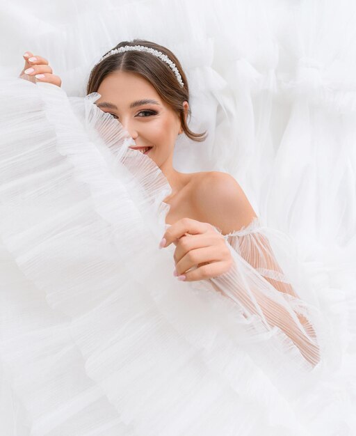 Felice bella giovane donna sdraiata sul suo matrimonio bianco vestito gonfio guardando nella fotocamera Sorridente sposa ragazza con perfetto trucco nudo e gioielli Abito da sposa concetto Sposa adorabile Salone di nozze