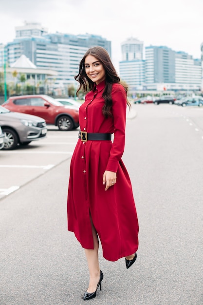 Felice bella giovane signora in abito rosso che guarda l'obbiettivo mentre si cammina lungo la strada nella città moderna. Concetto di stile di vita