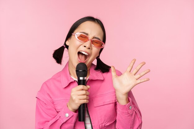 Felice bella ragazza asiatica che canta con il microfono utilizzando il microfono godendo il karaoke in posa su sfondo rosa studio