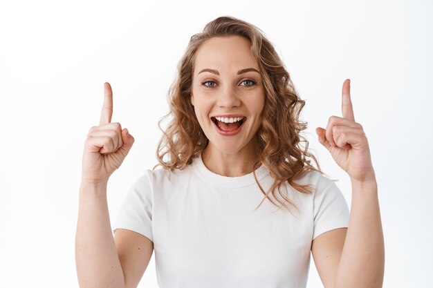 Felice bella ragazza con i capelli biondi ricci, puntando le dita in alto e sorridendo compiaciuta, mostrando pubblicità, in piedi sul muro bianco