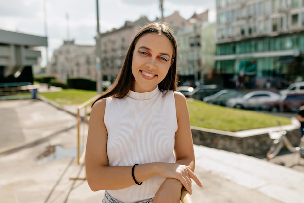 Felice bella ragazza con i capelli lunghi e trucco luminoso indossa una maglietta bianca sorridente alla telecamera e trascorrere del tempo all'aperto in una calda giornata di sole