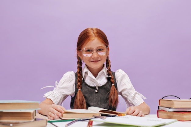 Felice bella ragazza con i capelli rossi in occhiali trasparenti e camicia bianca della scuola che esamina la macchina fotografica e sorridente su sfondo viola