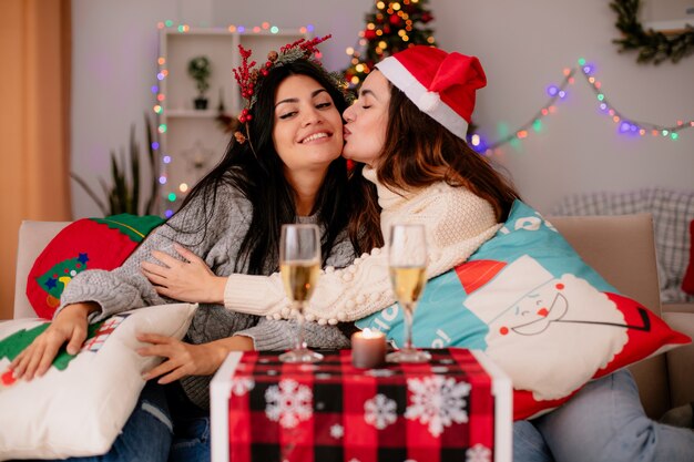 Felice bella ragazza con il cappello della santa bacia la sua amica con la ghirlanda di agrifoglio che si siede sulle poltrone e si gode il periodo natalizio a casa