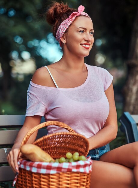 Felice bella rossa femmina che indossa abiti casual seduto con un cestino da picnic su una panchina in un parco.