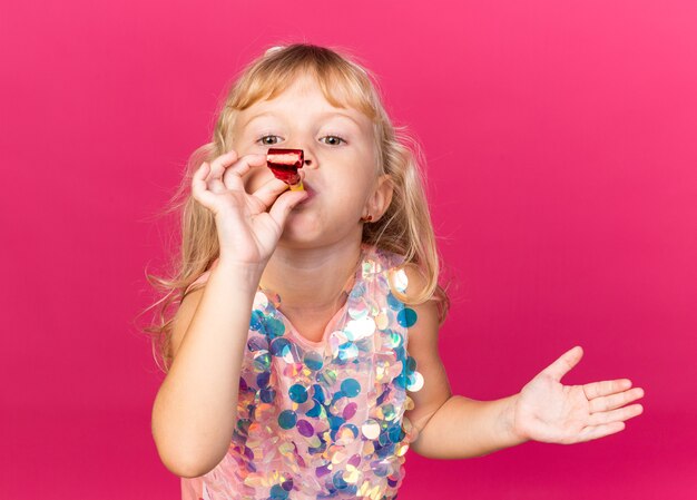 Felice bimba bionda che soffia fischio di festa isolato sulla parete rosa con spazio di copia