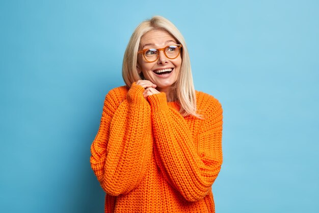 Felice bionda donna europea senior ricorda qualcosa di piacevole risate felicemente e tiene le mani premute insieme indossa occhiali da vista maglione arancione lavorato a maglia sciolto.