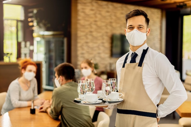 Felice cameriere che serve caffè ai suoi clienti mentre indossa una maschera protettiva in un bar