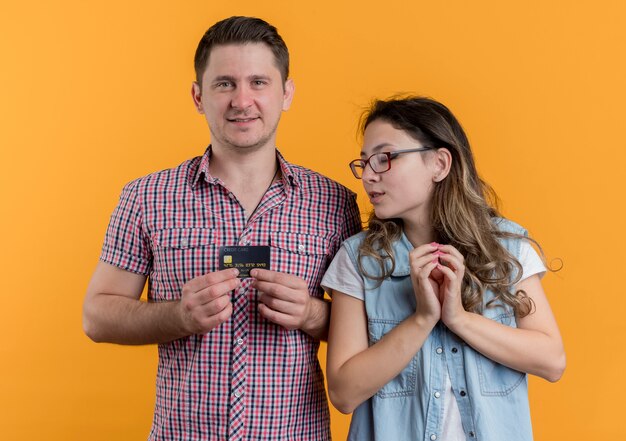 Felice coppia giovane uomo e donna utilizzando gadget donna che guarda lo schermo del suo fidanzato smartphone in piedi sopra la parete arancione
