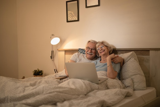 Felice coppia senior sdraiata a letto e navigare in rete sul laptop di notte