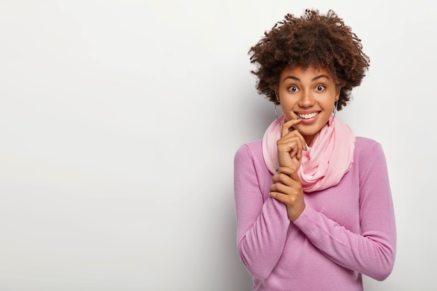 Felice donna afro con l'acconciatura riccia, tocca il dito indice sulle labbra, ha un sorriso a trentadue denti, vestita casualmente