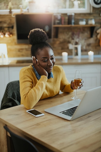 Felice donna afroamericana che usa il laptop mentre si frequenta online e beve champagne a casa