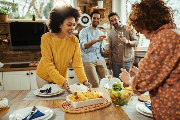 Felice donna afroamericana e la sua amica che servono cibo al tavolo da pranzo mentre i loro fidanzati bevono vino in background.