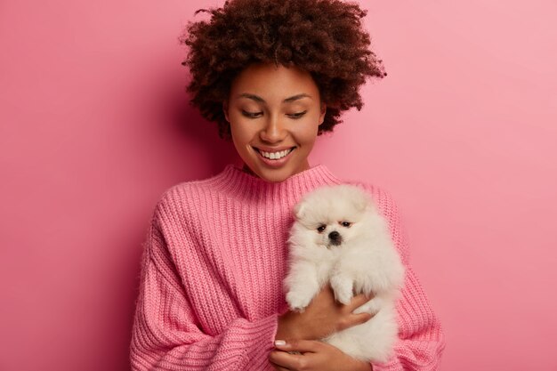 Felice donna afroamericana guarda con gioia il suo nuovo animale domestico, tiene in mano il cane spitz bianco, indossa un maglione oversize, sorride ampiamente, isolato su sfondo rosa.