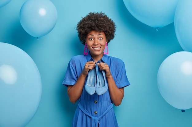 Felice donna afroamericana vestita in abito blu, tiene belle scarpe, abiti per occasioni speciali, si prepara per l'appuntamento serale. Felice donna di compleanno riceve le scarpe come regalo, posa vicino a palloncini blu